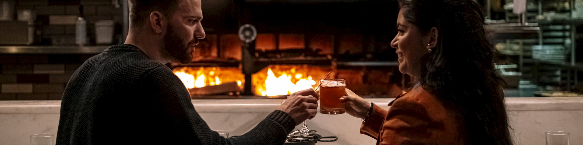 A couple is toasting drinks at a restaurant counter, with a brick oven burning brightly in the background.