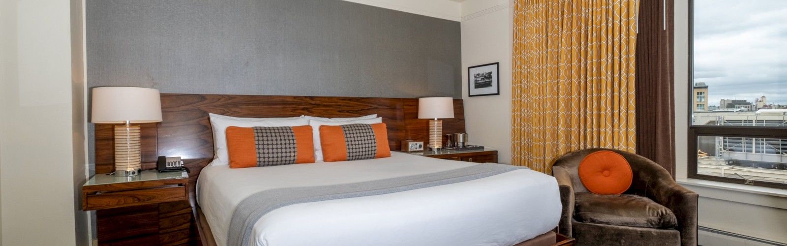 A hotel room with a king-sized bed, orange accents, nightstands, lamps, an upholstered chair, and a window with a city view in the image.