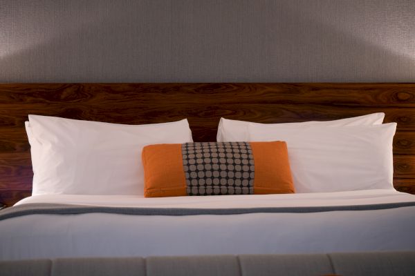 A neatly made bed with white sheets, two white pillows, an orange and patterned lumbar pillow, and a wooden headboard.