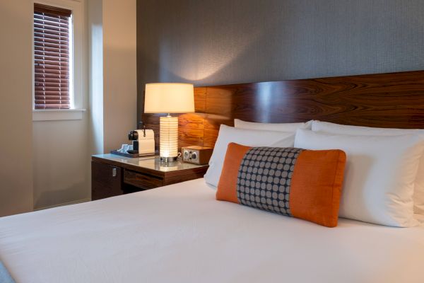 A cozy hotel room features a neatly made bed with white linens and an orange pillow, a nightstand with a lamp, and a coffee maker in the corner.