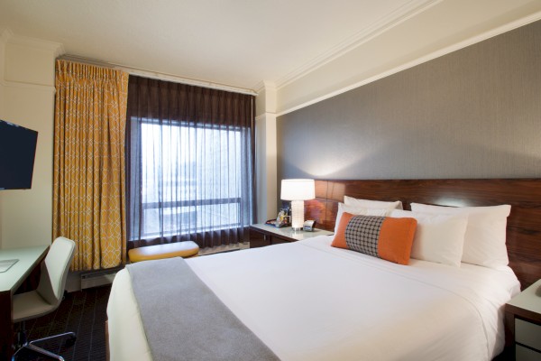 A hotel room with a neatly made bed, a desk, chair, and a window with curtains. Warm lighting and a TV are also present.