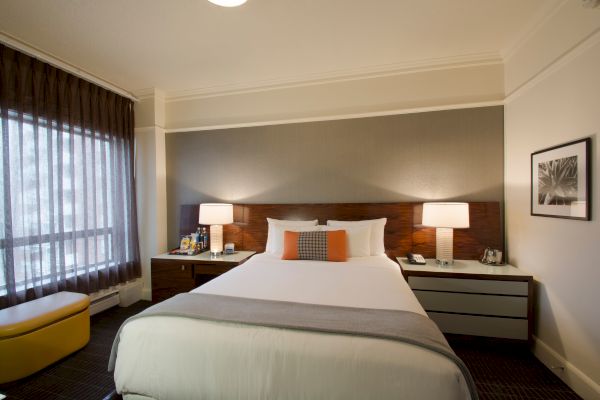 A cozy hotel room features a neatly made bed with a headboard, bedside lamps, two nightstands with items, a window with curtains, and a framed picture.