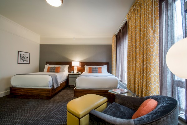 The image shows a hotel room with two beds, a lamp, patterned curtains, a chair with an orange cushion, and an ottoman by a large window.