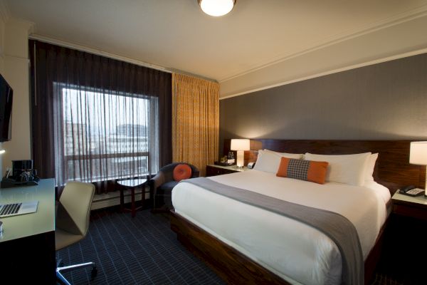A modern hotel room with a large bed, desk, chair, and window with curtains, decorated in neutral tones and orange accents, ending the sentence.