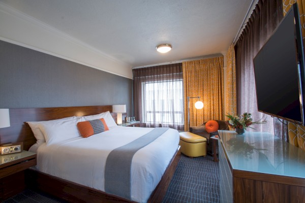 A modern hotel room with a king-sized bed, gray and orange decor, a flatscreen TV, and a small seating area by the window.