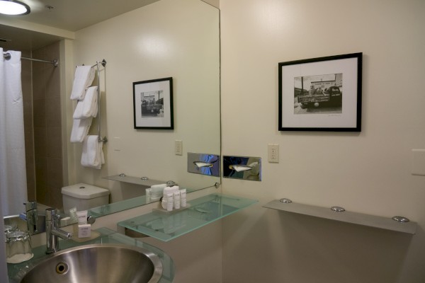 The image shows a modern bathroom with a round sink, mirror, towels, framed pictures, and toiletries on a glass shelf.