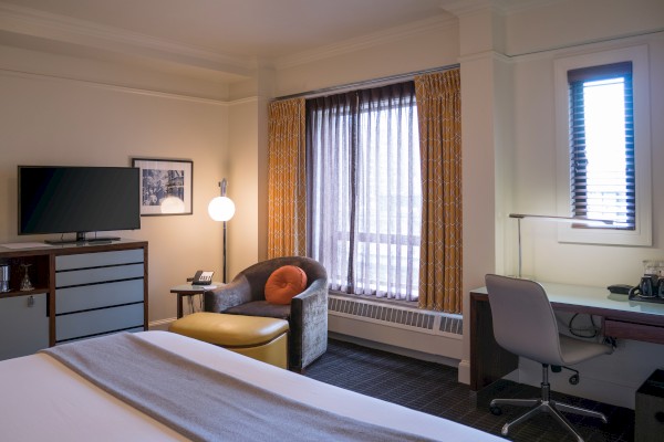 The image shows a cozy hotel room with a bed, TV, armchair, desk with chair, and window with curtains. A lamp and framed picture are present.