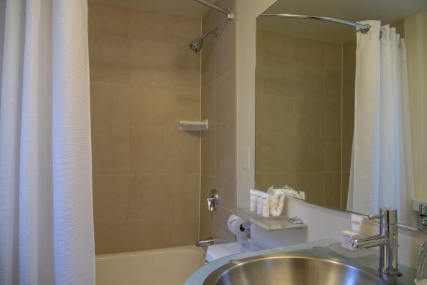 The image shows a bathroom with a shower, a sink, a mirror, a white shower curtain, and toiletries neatly placed on a shelf.