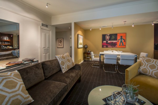 A modern living room with brown and yellow sofas, geometric pillows, a round table, and a dining area featuring abstract artwork on the walls.