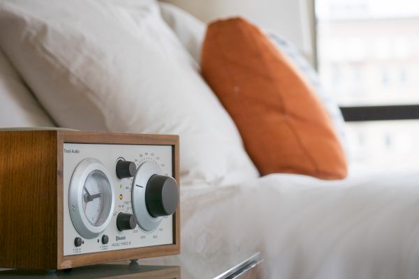 The image shows a wooden-cased radio on a bedside table with a white bed and an orange pillow in the background.