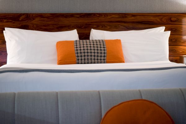 The image shows a neatly made bed with two white pillows, an orange and patterned cushion, and a headboard, suggesting a hotel or modern bedroom.