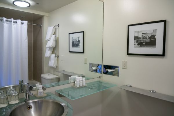 A modern bathroom with a stainless steel sink, glass countertop, toiletries, mirror, wall art, towels on a rack, toilet, and shower curtain ending the sentence.