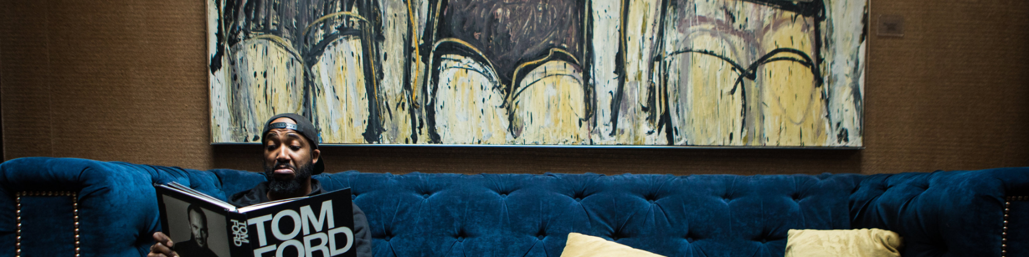 A man reads a Tom Ford book while sitting on a blue sofa with two yellow pillows, in front of an abstract painting of chairs.