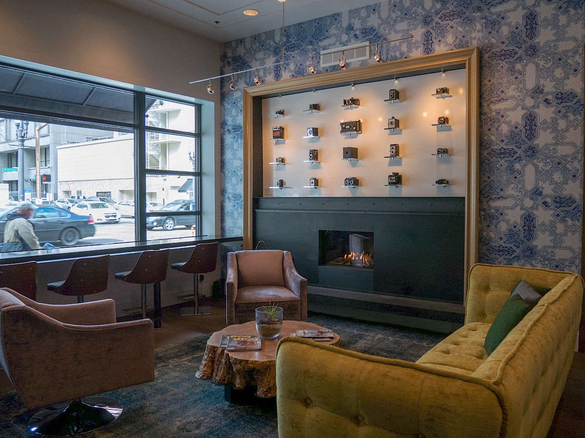 The image displays a cozy indoor seating area with modern furnishings, a fireplace, wall decor, and large windows overlooking a street scene.