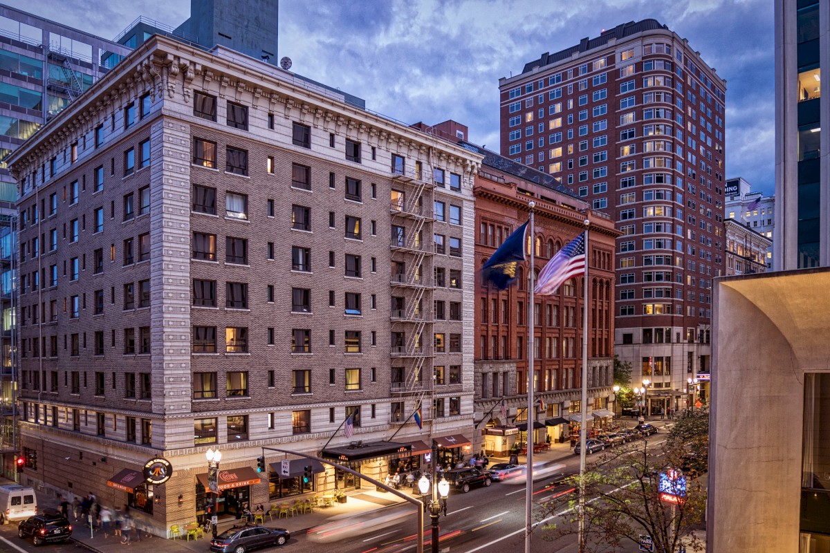 The image shows a city street with multi-story buildings, pedestrians, and vehicles. Flags are flying near the building's entrance.