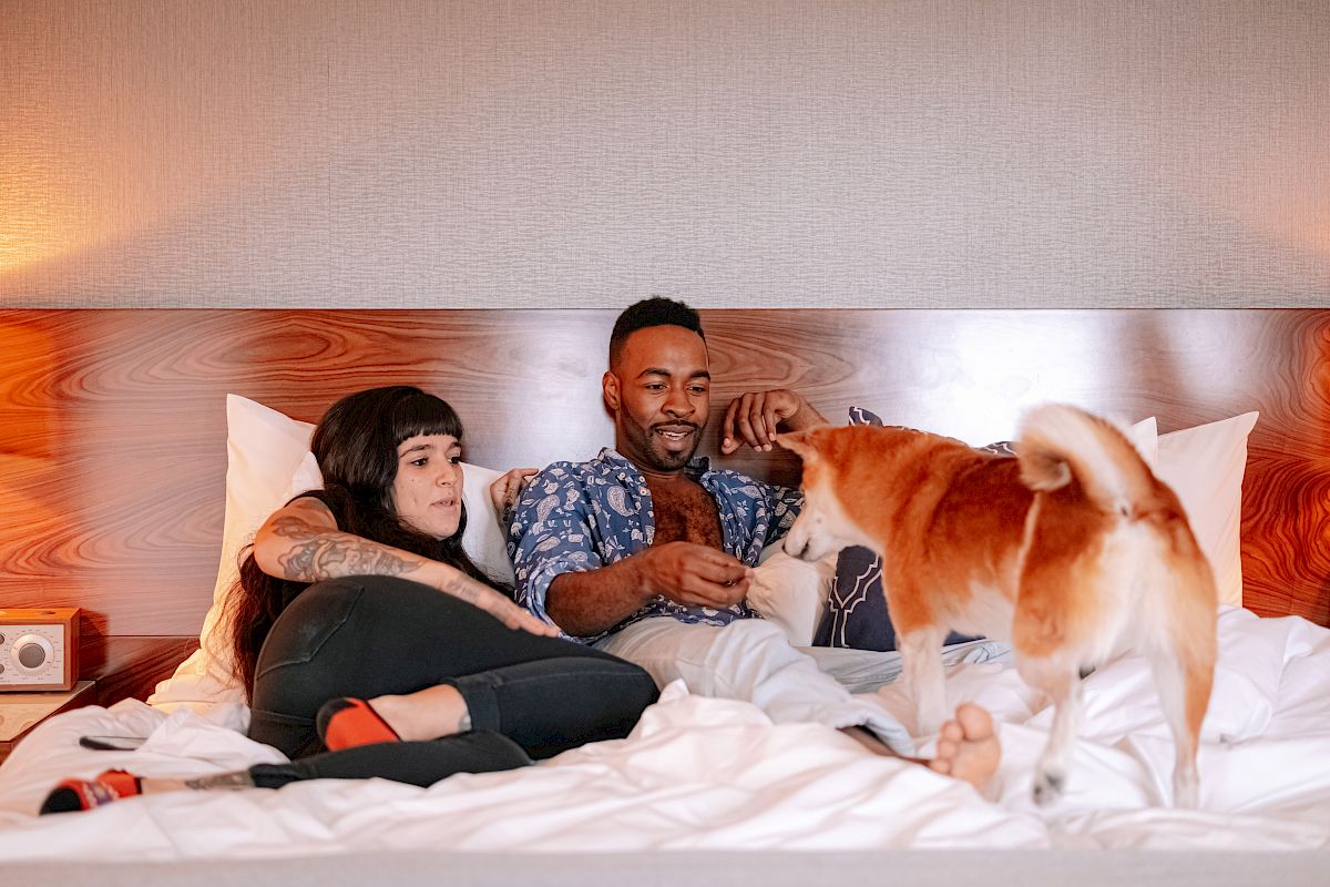 A couple relaxes on a bed while interacting with a dog; the woman is sitting up, and the man seems to be feeding the dog.
