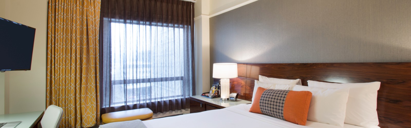The image shows a hotel room with a bed, bedside table, lamp, desk, chair, TV, large window with curtains, and modern decor.