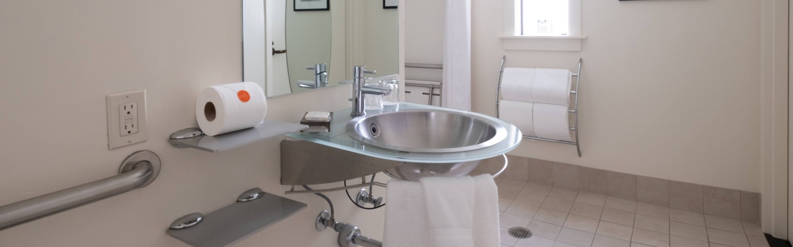 The image depicts a clean, modern bathroom with a sink, a roll of toilet paper, a towel, a shower, and two framed pictures on the walls.