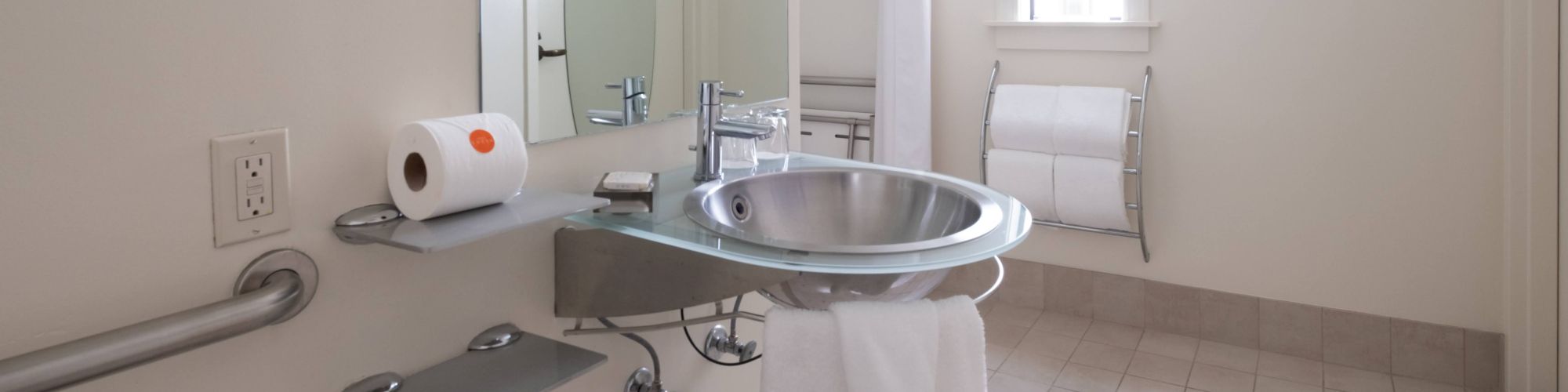 The image depicts a clean, modern bathroom with a sink, a roll of toilet paper, a towel, a shower, and two framed pictures on the walls.