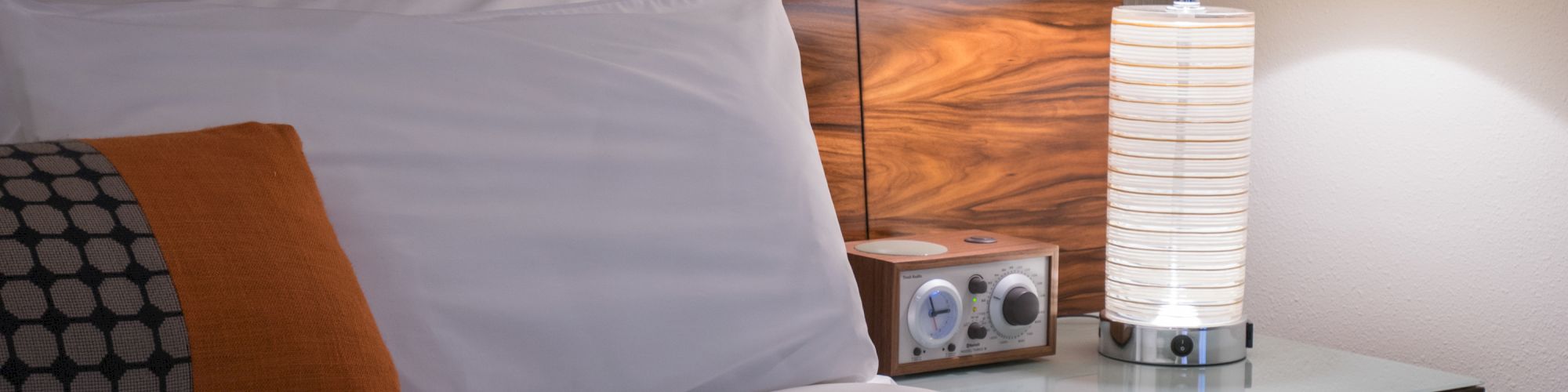 A neatly made bed with white sheets and pillows, orange and patterned cushions, beside a wooden nightstand with a lamp and clock radio.
