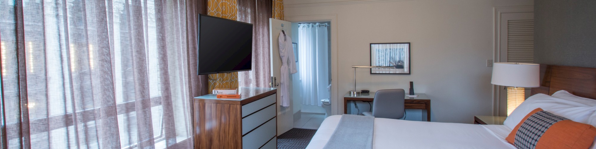 A cozy hotel room featuring a large bed, a small desk with a chair, a TV on a dresser, and sheer curtains covering a window.