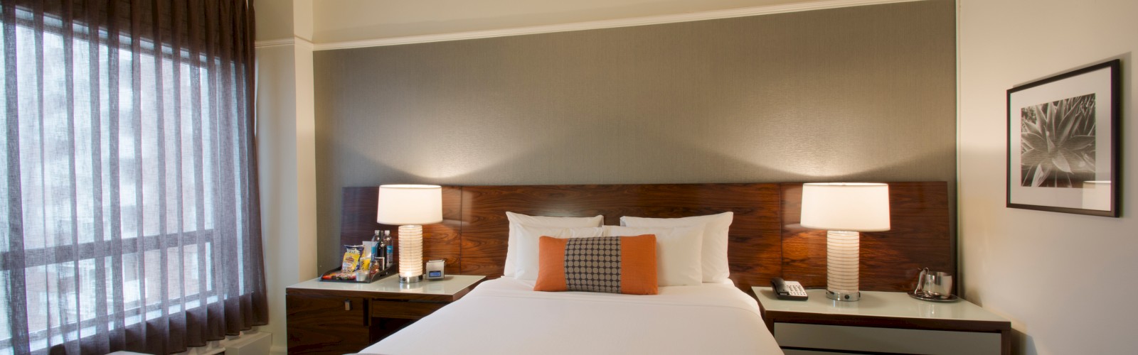 This image shows a modern hotel room with a neatly made bed, two bedside tables with lamps, a yellow ottoman, and a window with curtains.