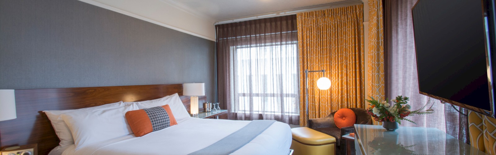 A modern hotel room with a large bed, decorative pillows, TV, table, and chair. The design features warm tones and ample natural light.