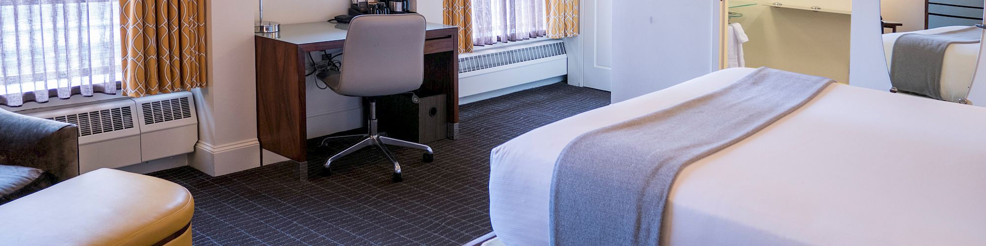 A bedroom with a bed, desk, chair, and large windows covered with curtains. There's an open door to a bathroom.