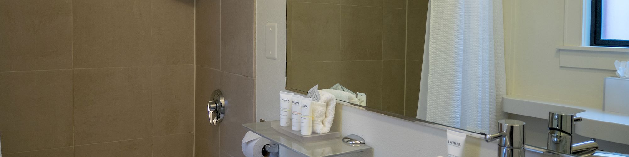 A clean, modern bathroom with a bathtub, white shower curtain, large mirror, sink, and neatly arranged toiletries on the counter.