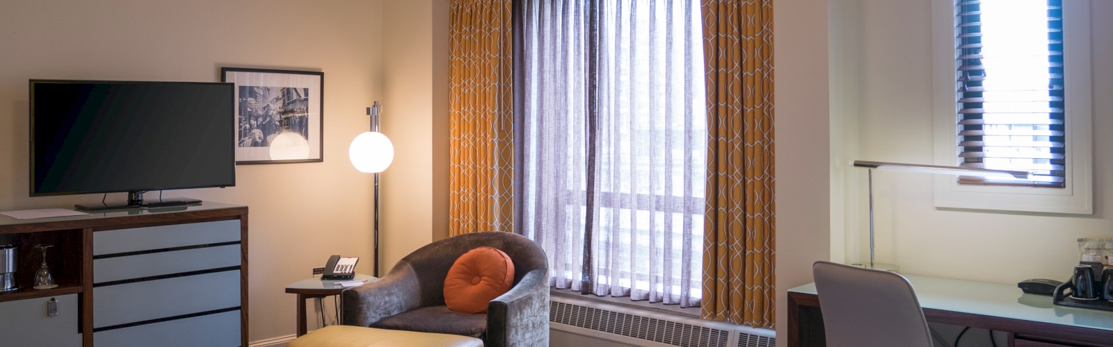 A hotel room with a bed, TV, chair, lamp, desk, and window with curtains. Modern décor creates a comfortable atmosphere.