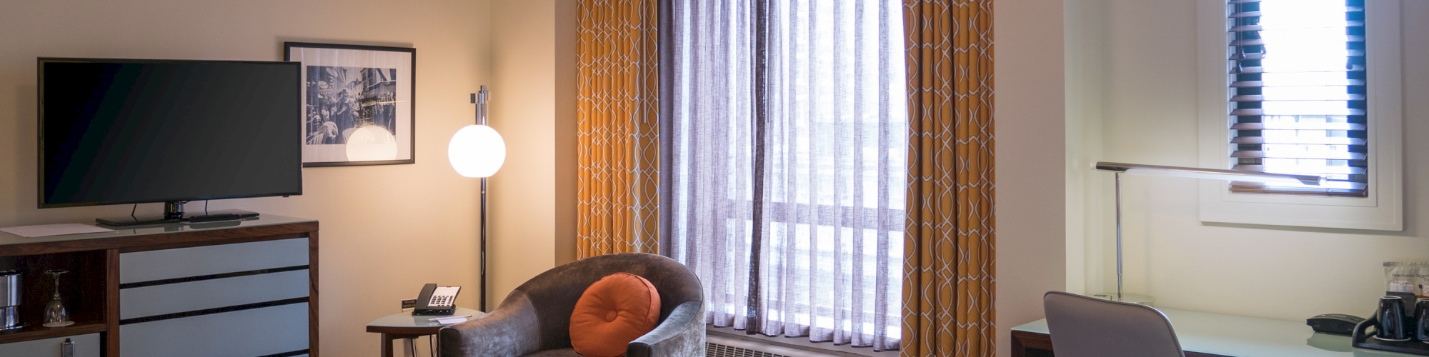 A hotel room with a bed, TV, chair, lamp, desk, and window with curtains. Modern décor creates a comfortable atmosphere.