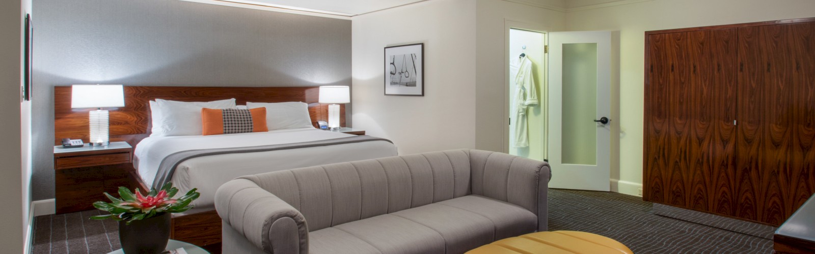 A spacious hotel room features a king-sized bed, a gray sofa, an orange ottoman, and a modern bathroom.