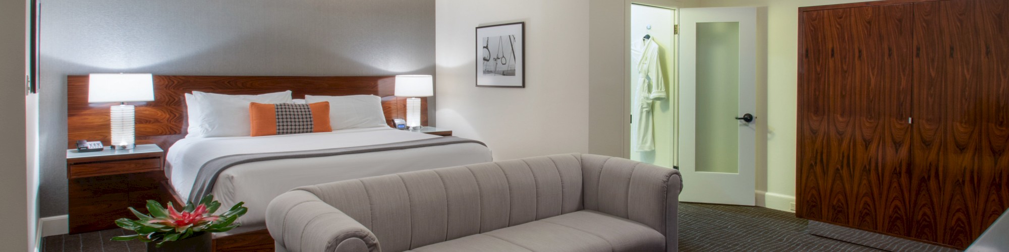 A spacious hotel room features a king-sized bed, a gray sofa, an orange ottoman, and a modern bathroom.