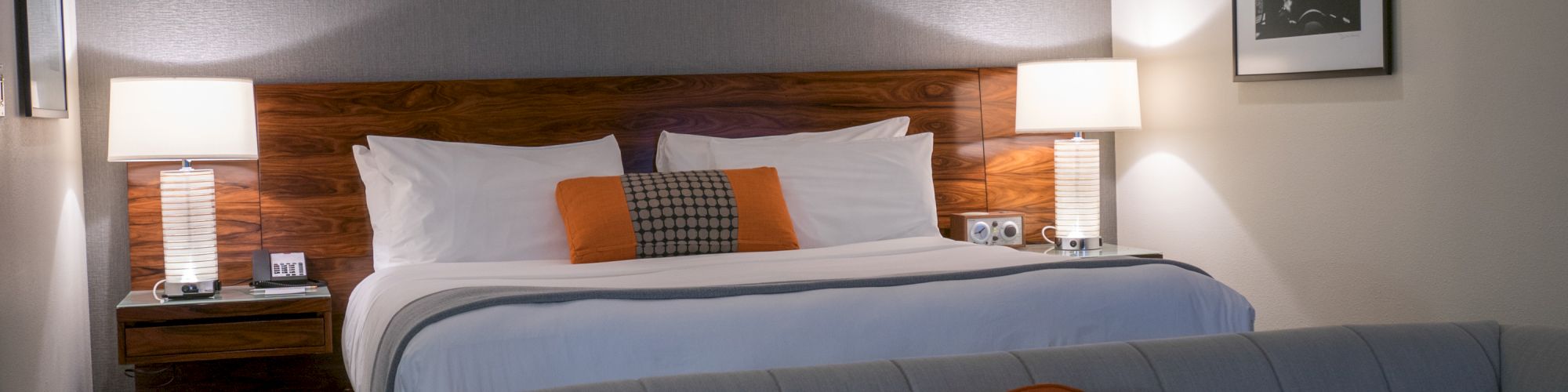 A cozy hotel room featuring a large bed with white linens and orange accents, flanked by two lamps, and a sofa with an orange cushion in the foreground.