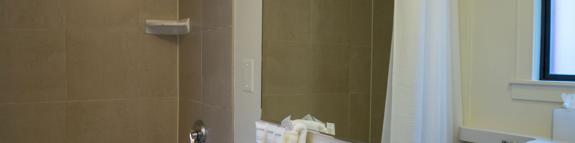 A bathroom with a sink, mirror, and shower. Toiletries are on the counter next to the sink. A glass and towel are also present.