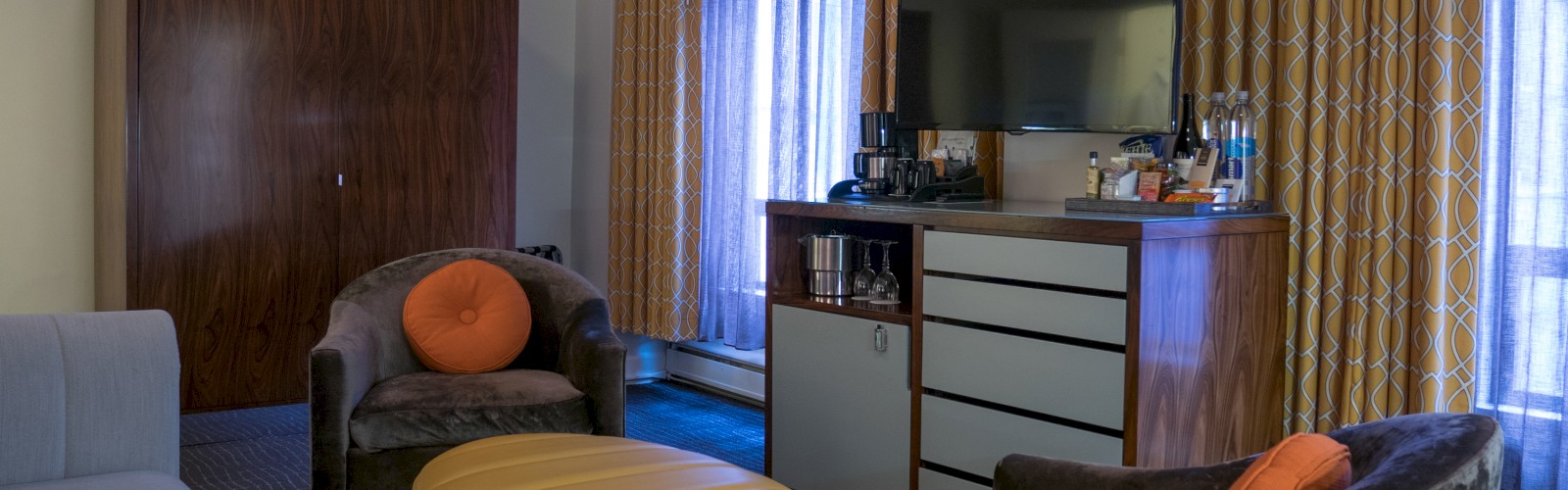 A cozy living room with chairs, a yellow ottoman, a TV, and a cabinet. Curtain-covered windows create a warm atmosphere.