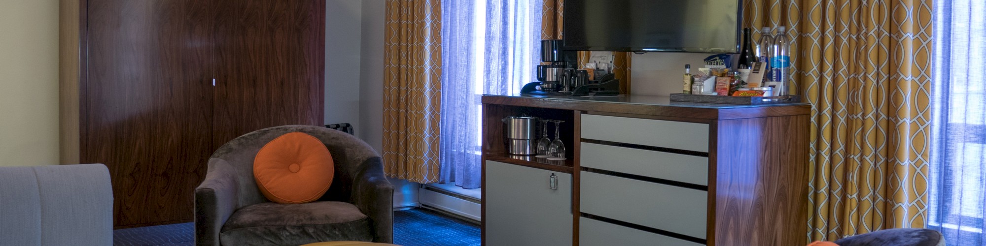 A cozy living room with chairs, a yellow ottoman, a TV, and a cabinet. Curtain-covered windows create a warm atmosphere.