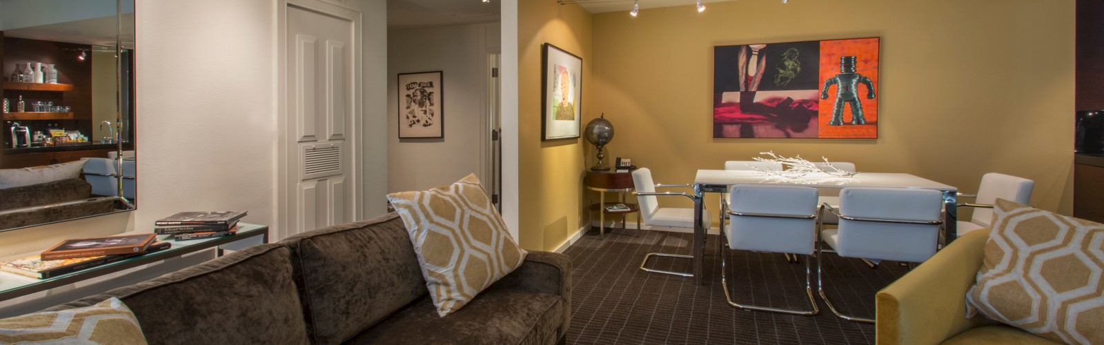 A cozy living room with a brown sofa, yellow pillows, dining area, vibrant art, and a modern decor style, creating a warm atmosphere.