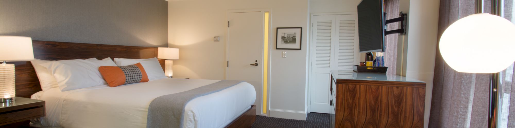 A modern hotel room with a bed, orange accents, a TV, a dresser, and seating area with an ottoman. A large window lets in natural light.