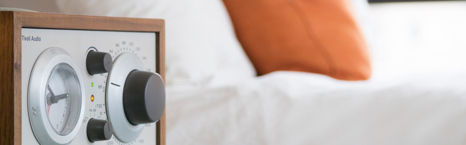 A retro-style radio with multiple dials sits on a bedside table next to a white bed with orange and white pillows, providing a cozy ambience.