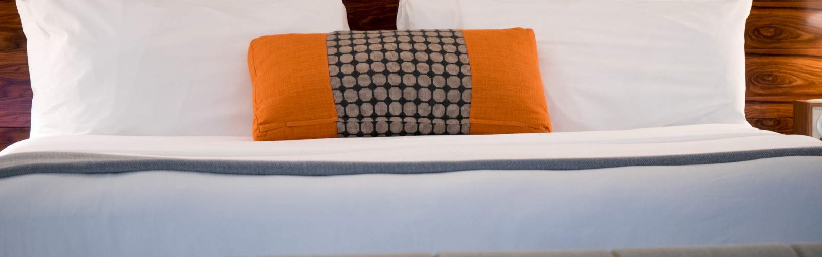 The image shows a neatly made bed with white pillows and an orange accent pillow with a pattern, set against a wooden headboard.