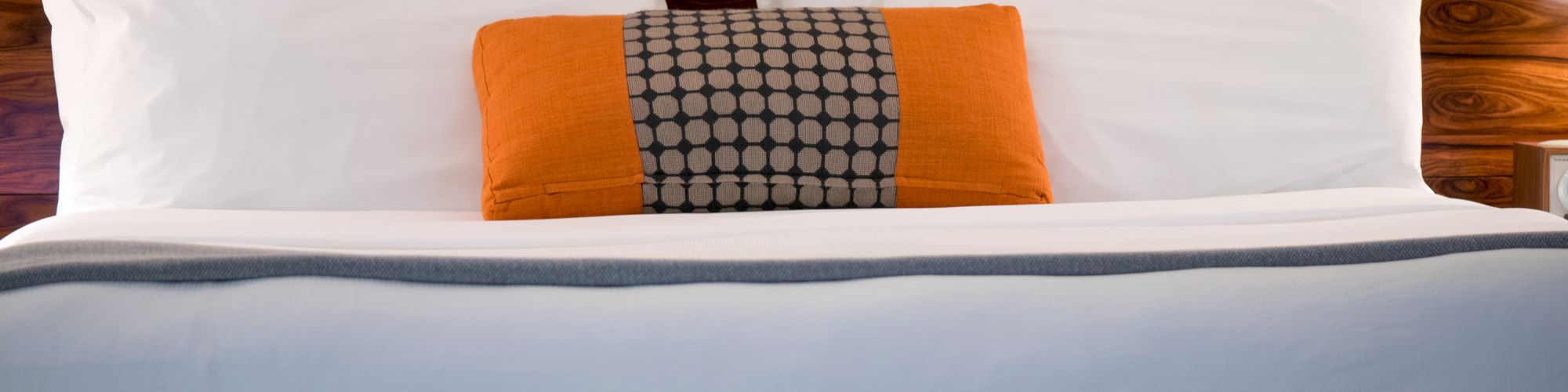 The image shows a neatly made bed with white pillows and an orange accent pillow with a pattern, set against a wooden headboard.