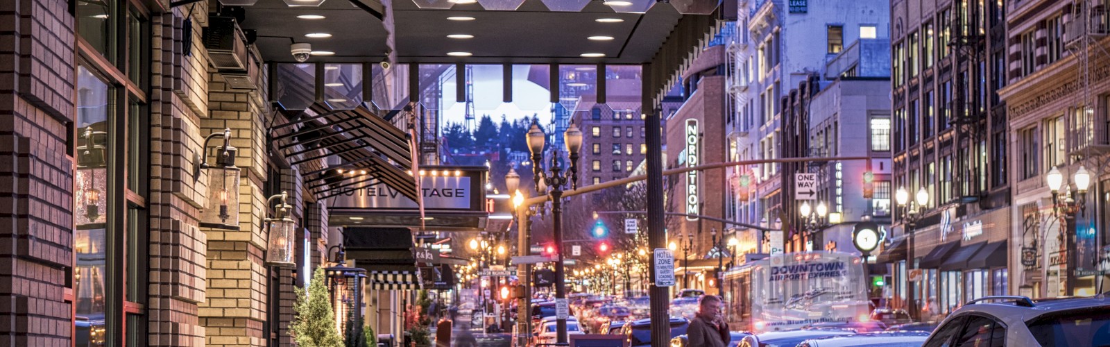 A lively urban street during dusk with Hotel Lucia's entrance visible, illuminated by streetlights and busy with pedestrians and cars.