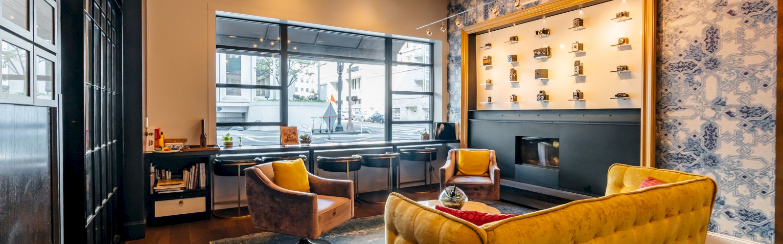 A stylish lounge with a yellow sofa, armchairs, art, shelves, large window, and modern chandelier on a cozy rug.