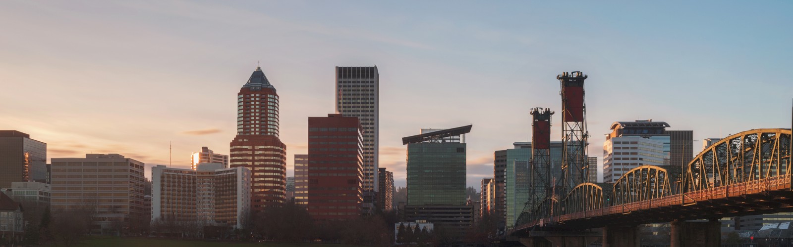 A city skyline at sunset with buildings reflecting in calm water and a bridge on the right side.