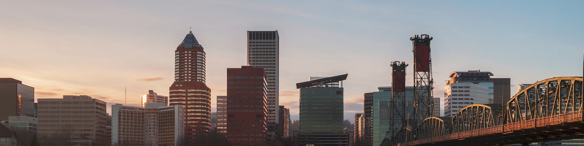 A city skyline at sunset with buildings reflecting in calm water and a bridge on the right side.