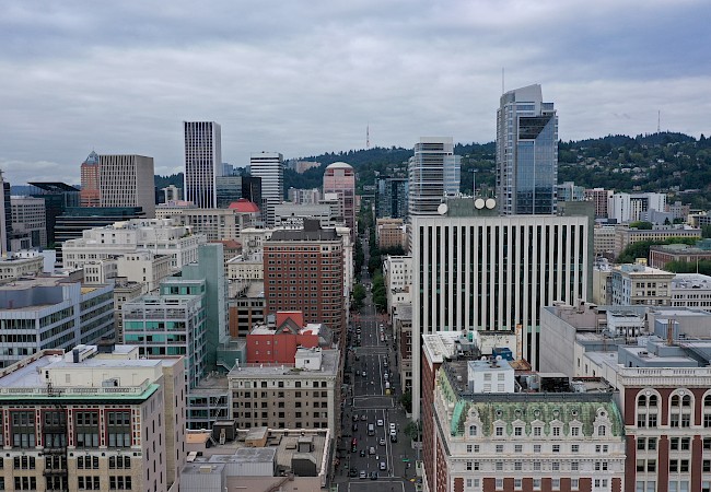 The image shows a cityscape with several skyscrapers, buildings, and a hilly landscape in the background under a cloudy sky.