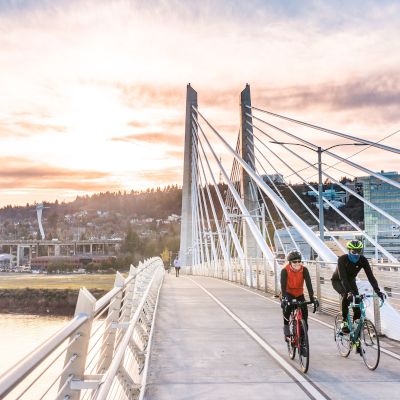 Two cyclists ride on a modern, cable-stayed bridge at sunset, with an urban backdrop and water below. The sky is partly cloudy.