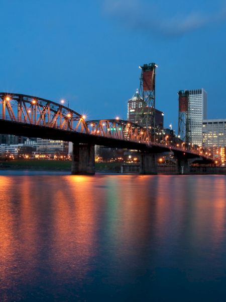 The image shows a lit-up bridge over a calm river with a city skyline in the background, captured during twilight, reflecting lights in the water.