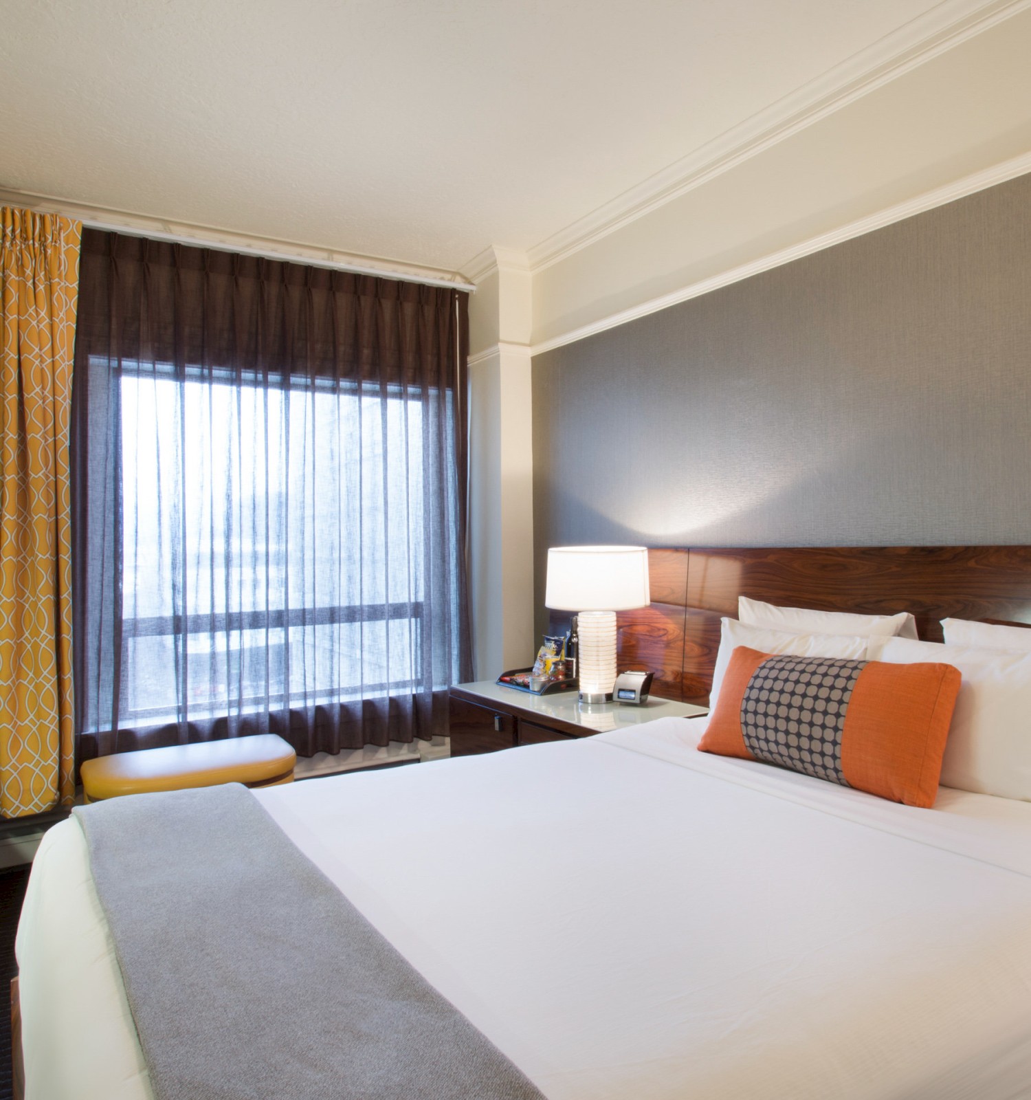 A modern hotel room with a neatly made bed, a TV, a desk chair, and large window with curtains. A lamp is on the bedside table.